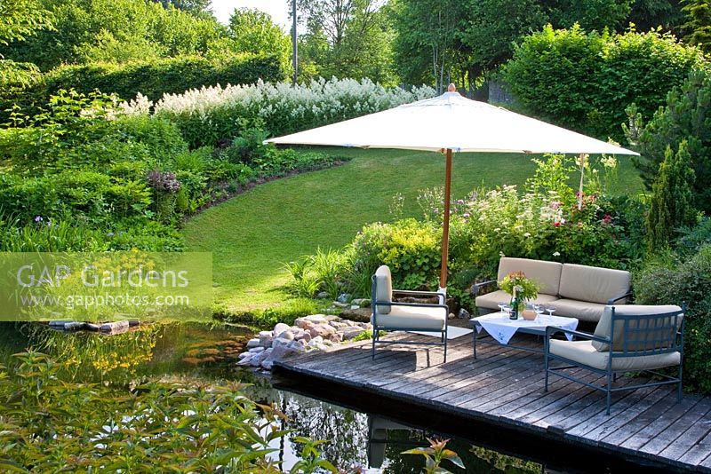 Relaxing area with parasol on wooden deck above a pond with primroses, flowering Aconogetum and a hedge encircle a lawn 