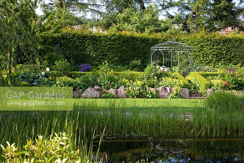 View over a natural swimming pool to a pavillon, backed with a hedge. The slope is secured by lime sand boulders, planting includes Alchemilla mollis, Buxus, Carpinus betulus, Fagus, Geranium, Hosta, Larix, Lupinus and Salix