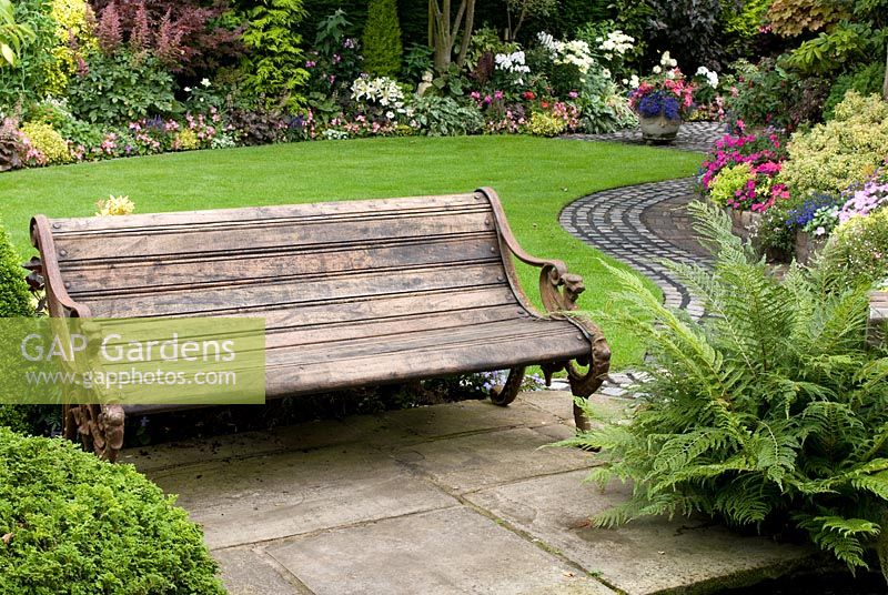 Old wooden bench in secluded, colourful garden with dense plantings of shrubs, conifers, ferns and bedding plants, path made from reclaimed slabs and cobbles - Brocklebank Road, Southport, Lancashire NGS
