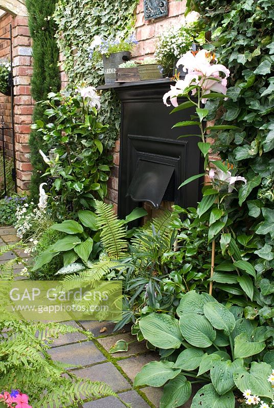 Reclaimed path of paviours and iron fireplace against red brick wall with Hosta, Lilium, Hedera and ferns - Brocklebank Road, Southport, Lancashire NGS