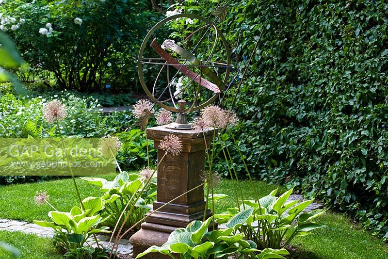 Sundial on a stone pillar backed by a hedge with Rosa 'Rambling Rector', flowerbed of Hosta fortunei 'Aureomarginata' and Allium giganteum - The Manor House, Germany
