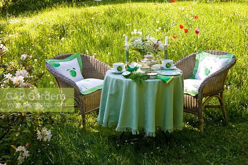 Table decorated with white china, candles and a bunch of wildflowers and two wicker armchairs in a meadow