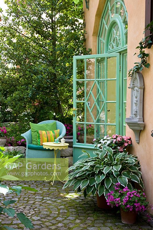 Turquoise wicker chair and yellow bistro table on paved patio with plants in terracotta pots next to classical styled glass door - Hosta, Ilex aquifolium, Impatiens and Oxalis