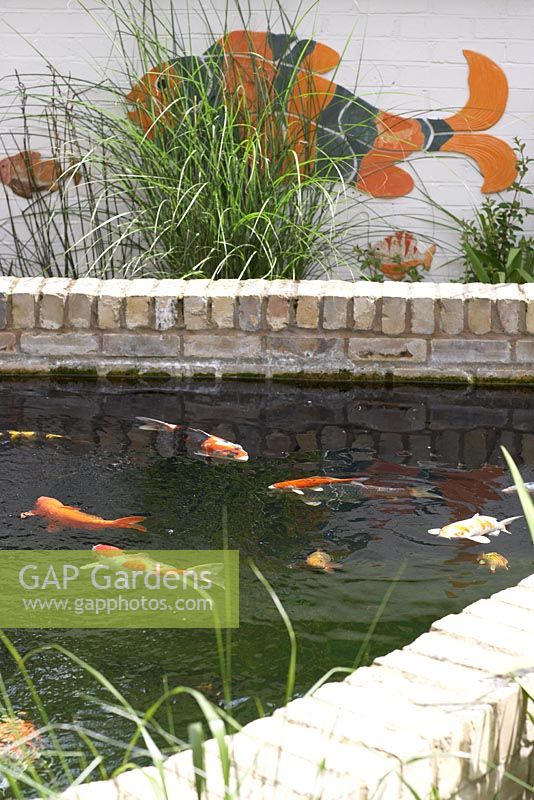 City garden showing pond with fish, with fish mural and ornamental grasses behind.