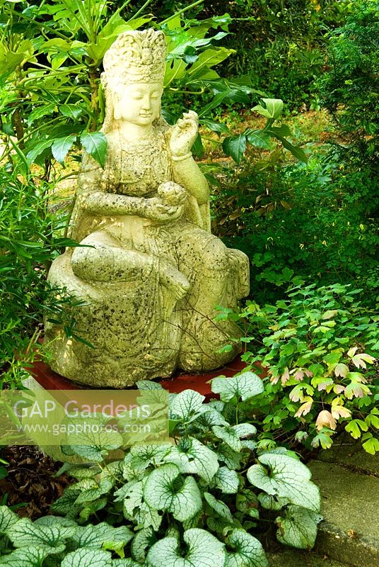 Chinese buddha surrounded by foliage plants including Brunnera, Choisya and Fatsia. Beggars Knoll, Newtown, Westbury, Wiltshire, UK
