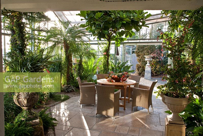 View into the glasshouse with tropical climate, wicker chairs and wooden table with a fruit arrangement. Planting includes Anthurium andreanum, Araucaria heterophylla, Beaucarnea recurvata, Billbergia nutans, Citrus sinensis, Cycas revoluta and Phoenix roebelinii - Wintergarten, Germany
