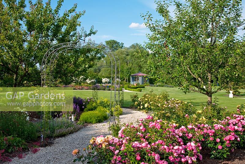 Metal arch over a gravel path leading towards a blue pavilion in a country garden with a large lawn and fruit trees. Borders of Rosa 'Guirlande d'Amour', Alchemilla mollis, Coreopsis verticillata 'Zagreb', Malus and Salvia nemorosa
