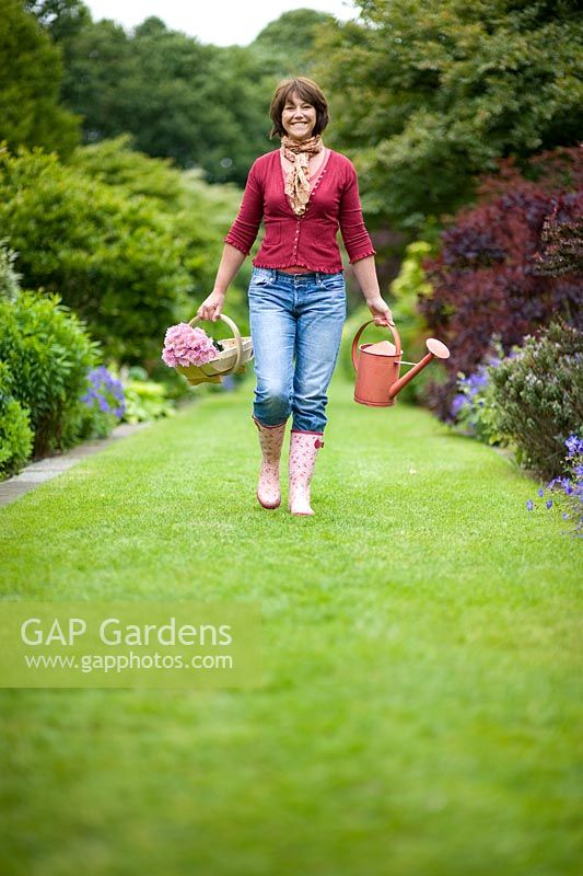 Woman wearing blue jeans and wellies carrying a red watering can and a wooden trug of pink chrysanthemum