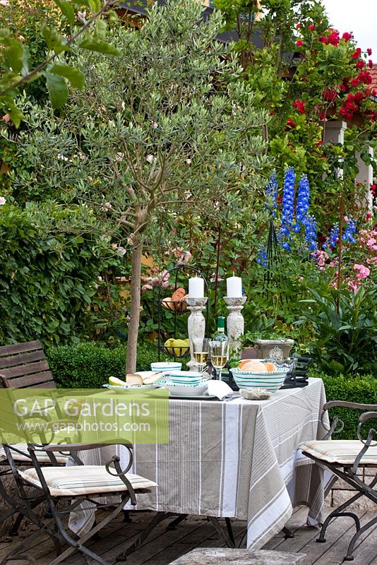 A linen dressed table with stone chandeliers and garden chairs against the background of Olea europaea and Delphinium Giant-Gruppe 'Blue Bird Pacific'