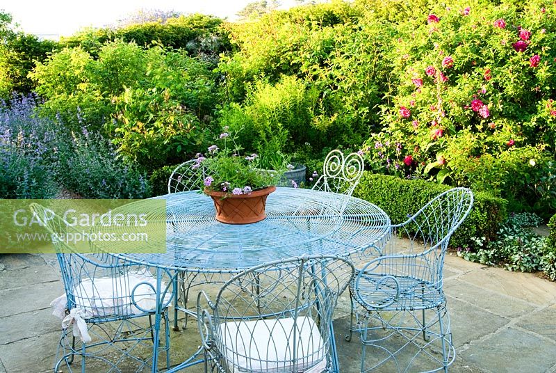 Decorative wirework chairs and table on the terrace outside the kitchen, with Nepeta 'Six Hills Giant' - Catmint, edging the path leading into the garden, and deep red Rosa 'Roseraie de l'Hay' - Old Rectory, Kingston, Isle of Wight, Hants, UK