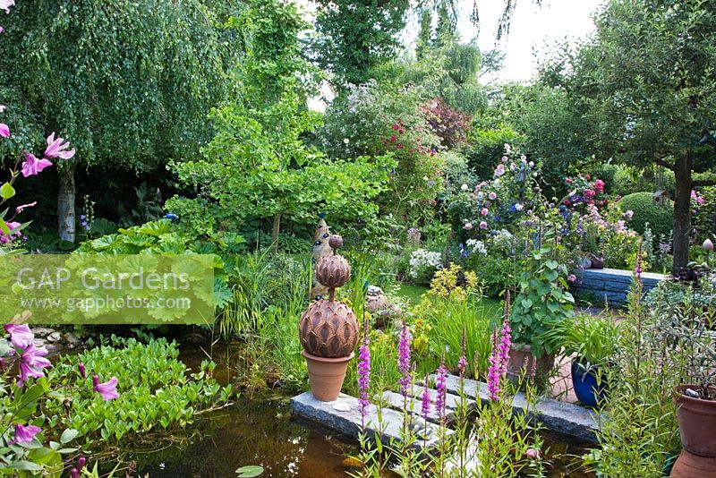 Plants and ceramic objects on stepping stones of a pond in a garden. Rosa 'Rosenresli', Darmera and Ginkgo biloba