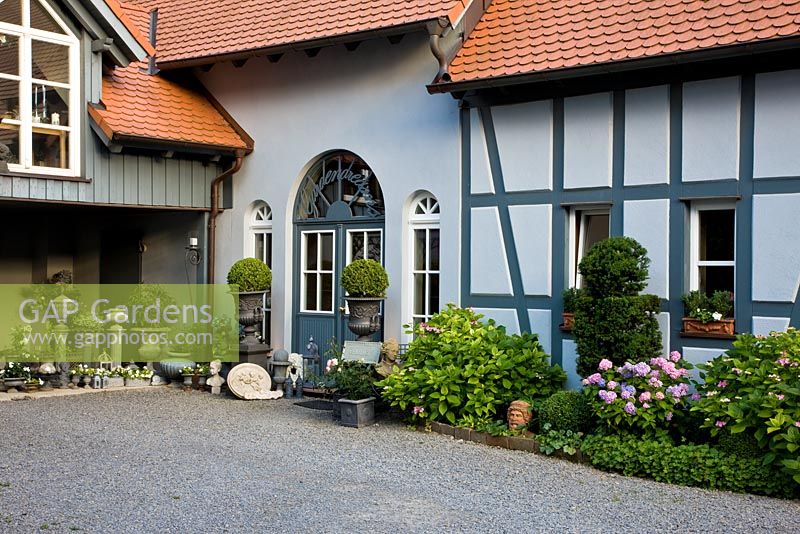 Entrance of timber framed house with decorative garden objects and urns on pillars. Buxus topiary, Geranium, Hydrangea macrophylla, Hydrangea serrata, Taxus 