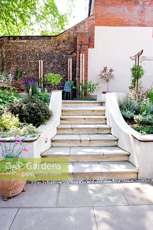 Steps through sloping small modern garden with white painted walls, terrace and tiered planting. 
