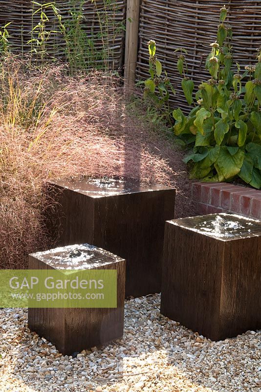 Wooden cube water features in gravel next to low brick wall and woven screen. Epimedium x versicolor 'Sulphureum', Anemanthele lessoniana, Phlomis russeliana