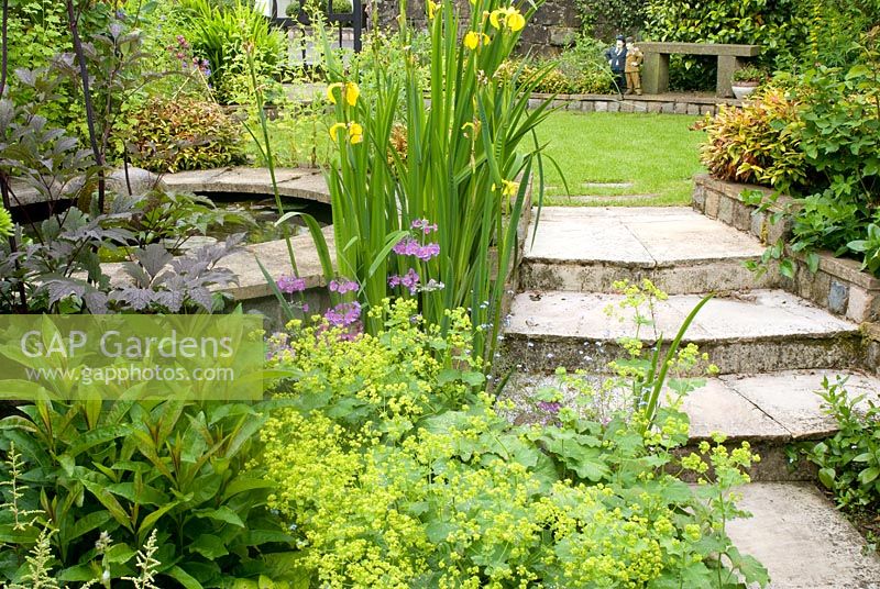 Stone path and steps leading to raised circular stone edged pond with adjacent lawn and perennials 