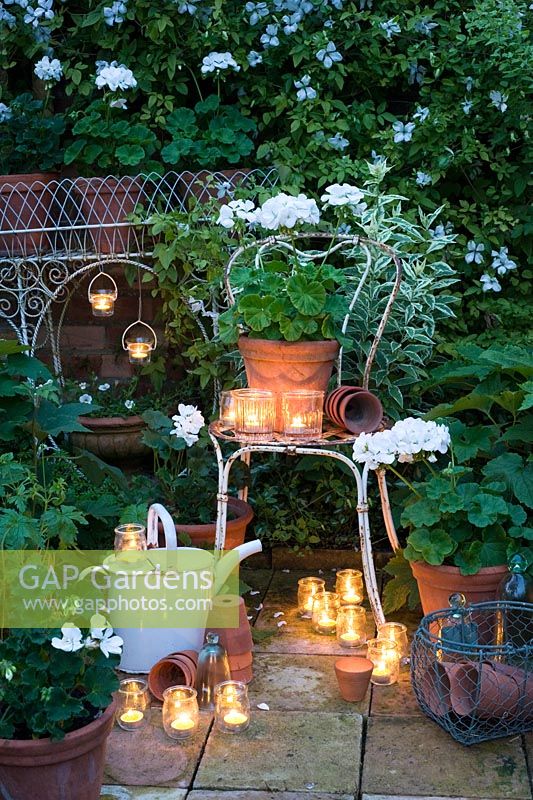 Glass jar tealights lighting and white geraniums in containers in white garden