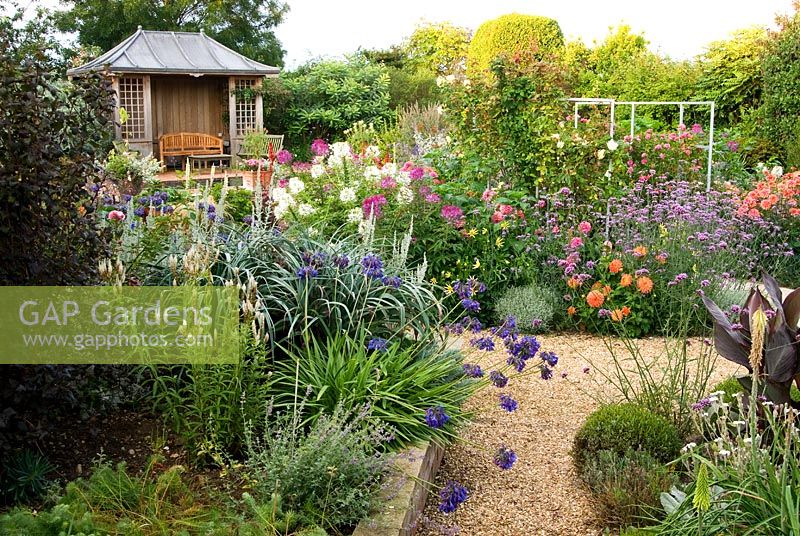 Central pond and summerhouse feature in the sunken garden, constructed in 2005, surrounded by a colourful mass of perennials and annuals including Eucomis, Verbena bonariensis, Dahlias, Agapanthus, Cleomes and dark leaved Cannas with gravel paths giving access - Isle of Wight, UK