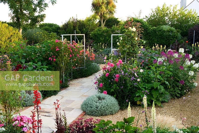 Sunken garden has a central stone path that passes between a metal frame supporting climbing Roses, with a mass of colourful perennials and annuals all around including Dahlias, Cleomes, Lobelia cardinalis and Verbena bonariensis - Isle of Wight, UK