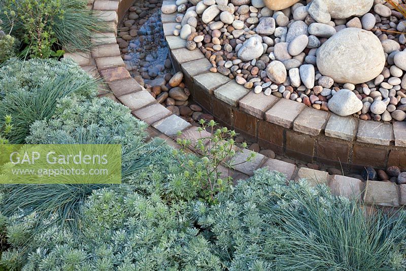 A circular rill of water between planting of silver foliage grasses, perennials, gravel, pebbles and brick 