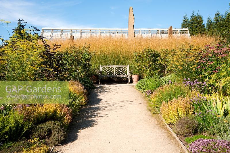 A bench in the herb garden with roof of Alpine House  beyond - RHS Garden Harlow Carr, Harrogate, North Yorkshire, UK