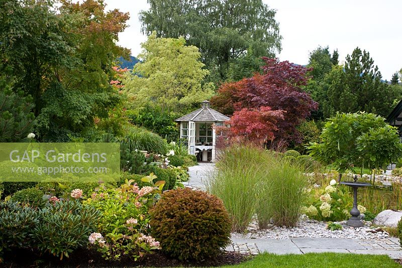 Amidst decorative shrubs and backed by large trees, a wooden pavilion with box spheres, flagged path and grasses. Planting includes Acer negundo 'Variegatum', Acer palmatum, Acer saccharinum, Betula pendula, Buxus, Hydrangea, Miscanthus sinensis, Pinus, Rhododendron, Taxus baccata and Wisteria