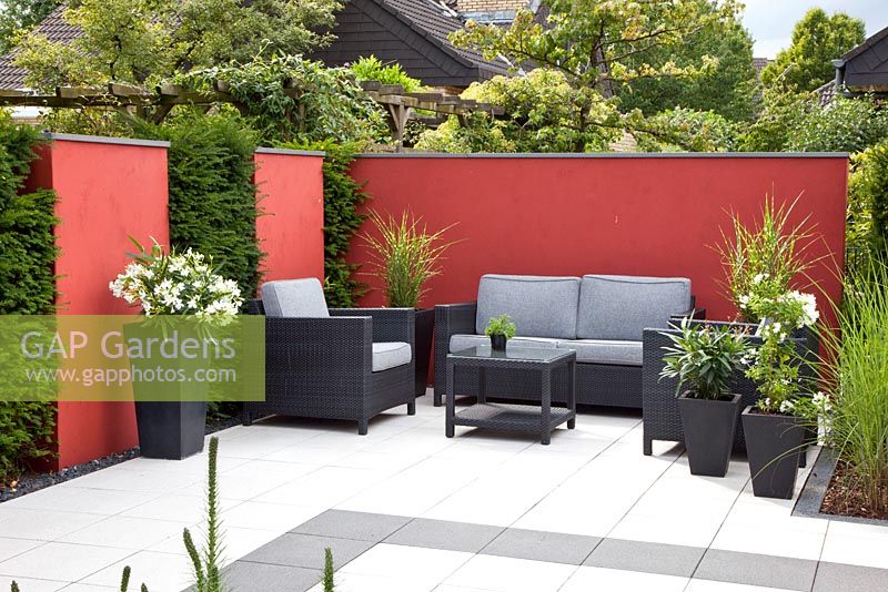 Modern patio with sofa and pots backed by red screen and Taxus - Yew hedge. Plants include Miscanthus and Nerium oleander