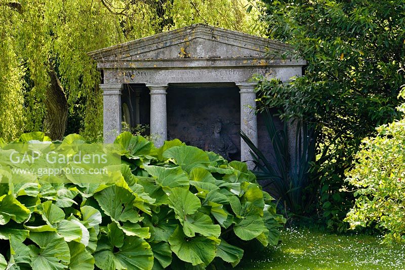 Temple in damp part of garden with Petasites alba - Butterburr in front. Salix x sepulcralis var. chrysocoma - Weeping willow behind temple. Statue of 'Architecture' inside temple