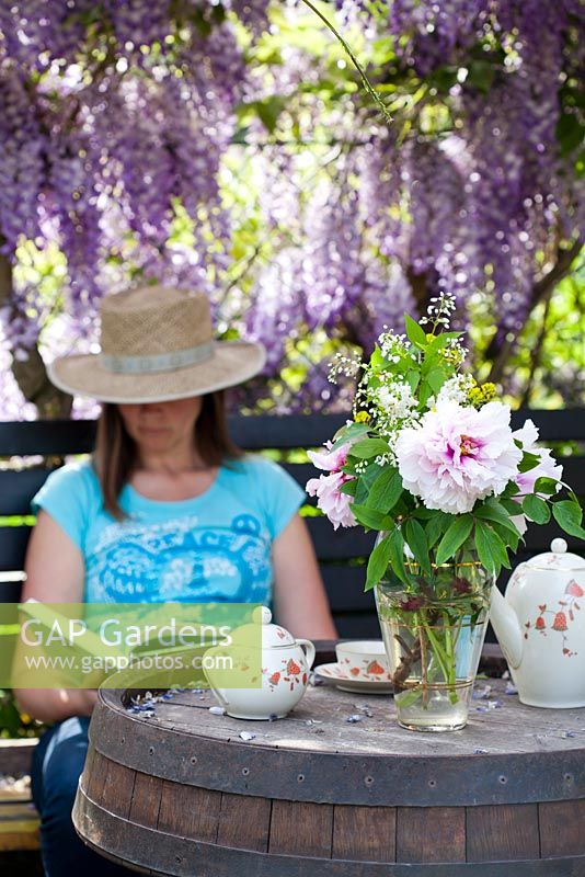 Woman reading a book in the shade under Wisteria, vase of Paeonia and Deutzia