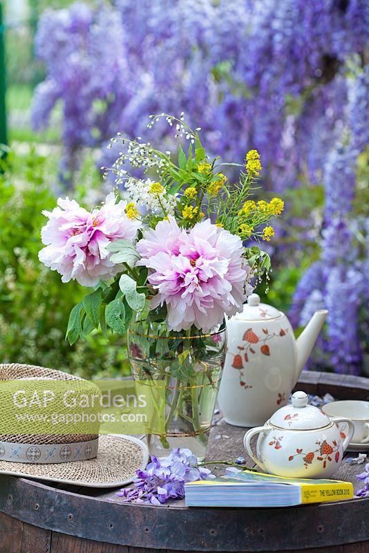 Vase of Paeonia and Deutzia with tea set, book and sun hat, Wisteria in background
