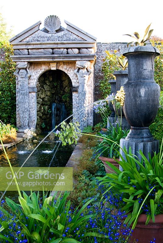 Rill pool flanked by turned oak urns spouting water through gilded lions' heads, with golden agaves in their tops. Water emerges from a rocky grotto framed by a scallop shell pediment and muscular caryatids. The Collector Earl's Garden designed by Julian and Isabel Bannerman.