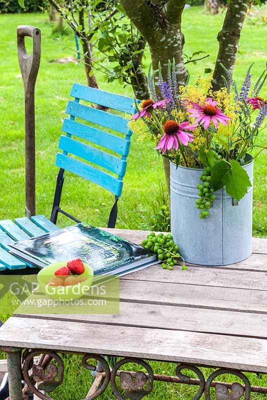 On a wooden table a vase with coneflower, Ladys Mantle, Veronica and grapes. Also on the table is a gardening book and a bowl of strawberries. In the background a colourful turquoise garden chair and a spade.