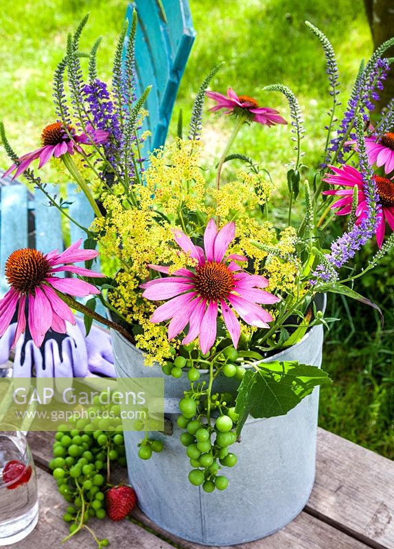 On a wooden table a vase with coneflower, Ladys Mantle and grapes. Also on the table is gardening gloves, hand fork and a glass of water. In the background a colourful turquoise garden chair.