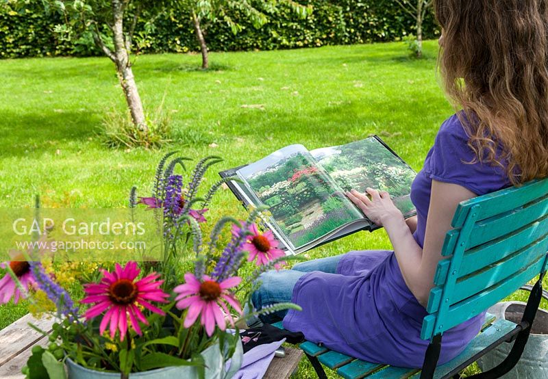 Woman wearing a purple summer dress sitting on a turquoise chair reading a gardening book in the shade.  In the foreground a vase with coneflower, Ladys Mantle and Veronica. On the wooden table gardening gloves.
