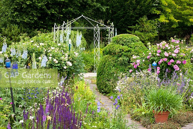 A paved pathway between flowering perennial borders and topiary leads towards a metal pavilion - Buxus, Campanula persicifolia, Delphinium Elatum-Grp, Nepeta faassenii and Salvia nemorosa - Germany