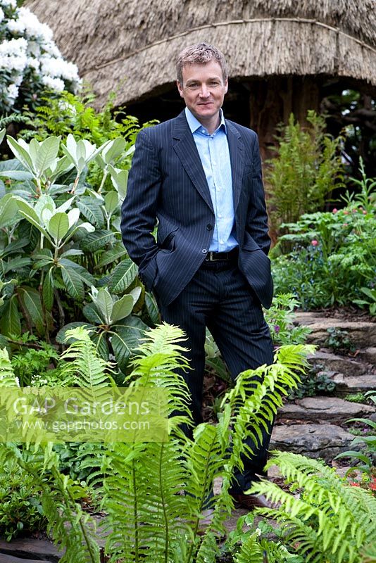 Portrait of Chris Beardshaw in Furzey Garden. Fern in foreground is Matteucia struthiopteris. Other plants include Rhododendron 'Hoppy', Geranium palmatum, Rhododendron 'Cunninghams White', and Primula 'Apple Blossom'.
