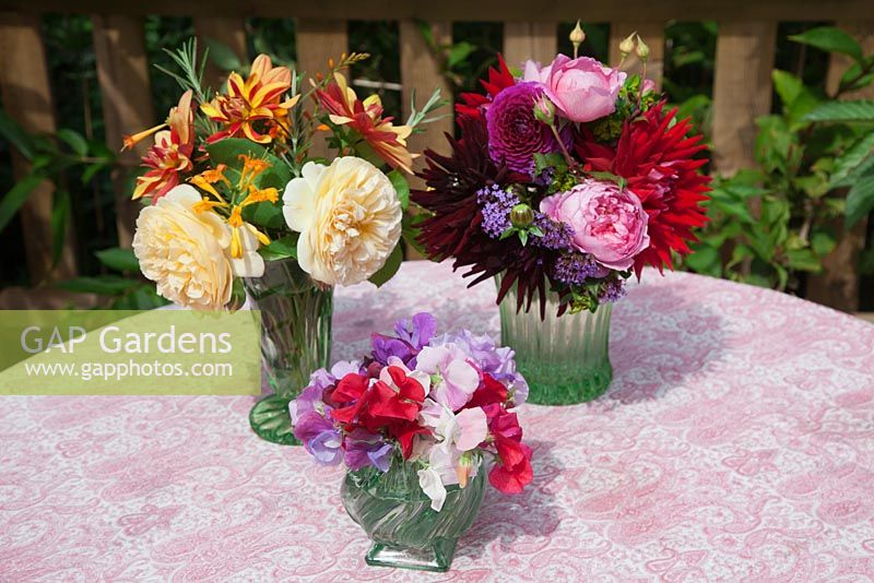 Flower arrangement with trio of Lathyrus latifolius - mixed Sweet Peas, mixed Dahlias and Verbena, Roses and Crocosmia in vintage glass vases on garden table.