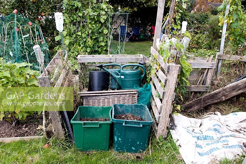 Neat, rustic fenced in storage area on allotment with compost in green recycling bins and plastic watering cans behind - Fortis Green Allotments, London Borough of Haringey