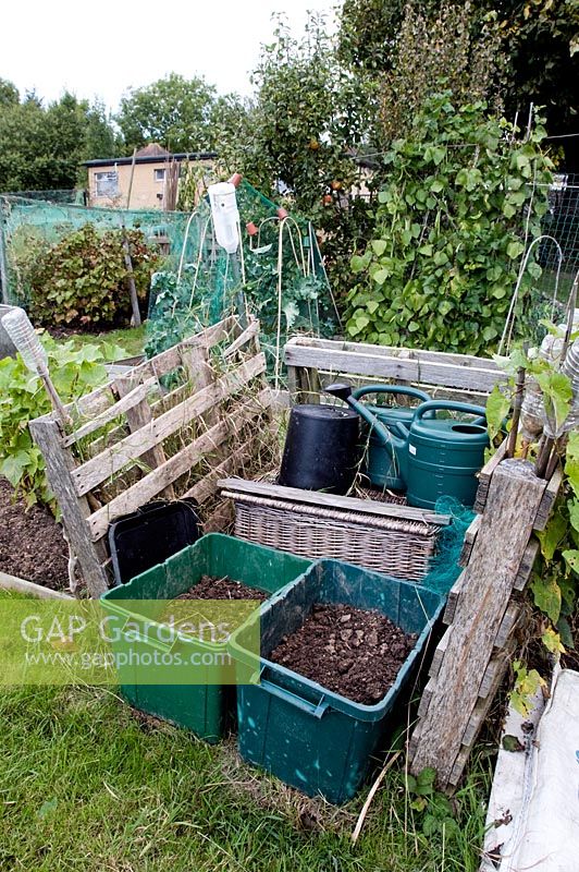 Neat, rustic fenced in storage area on allotment with compost in green recycling bins and plastic watering cans behind - Fortis Green Allotments, London Borough of Haringey 1