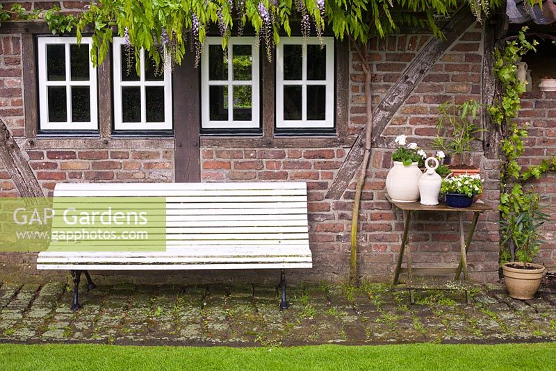 White painted wooden bench on a small patio by the summer house with Wisteria growing up a brick wall, De Carishof