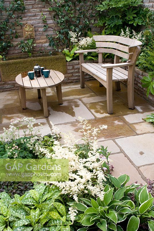 Wooden bench and table on stone patio with brick raised bed, water trough and fountain and planting of Astilbe, Ajuga, Astrantia, Pulmonaria, Hosta, Geranium, Hedera and ferns in the 'Enchantment' garden 