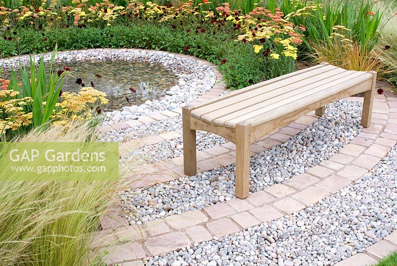 Wooden bench overlooking central rippling pool with spreading circles of pebbles, cobble setts and planting of Cosmos atrosanguineus, Achillea and Stipa tenuissima in the 'To The Beat' garden. Gold medal winner and Best Orchestra Garden - RHS Tatton Flower Show 2012