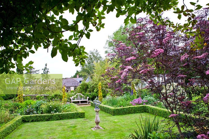 View of the garden with sundial in the centre of the lawn and Sambucus nigra 'Black Lace' in the foreground