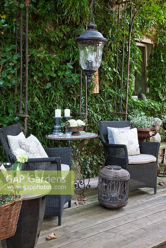 Modern styled black wicker chairs with white linen cushions on a wooden deck. Table with candle holders, a lantern and other decorative metal objects against a backdrop of Hedera helix