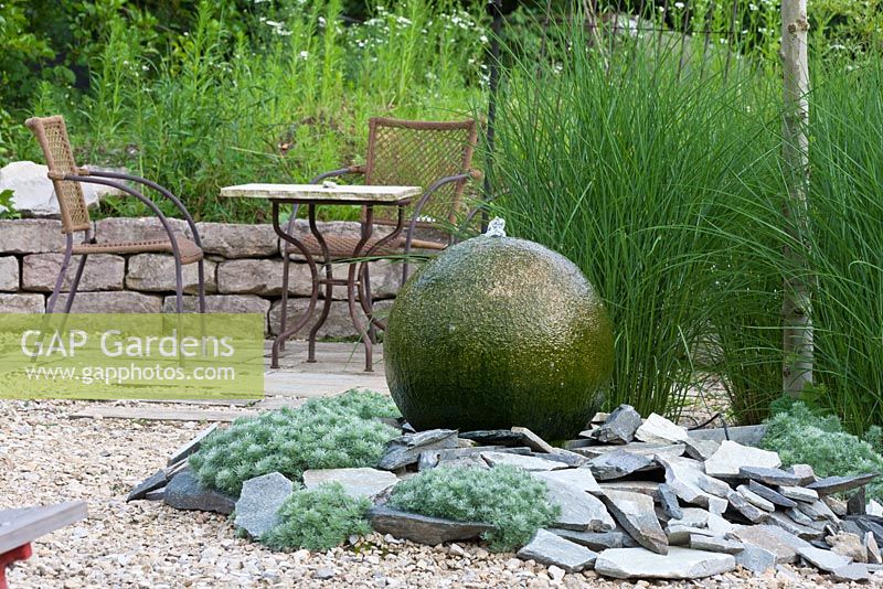 Water feature made of granite sphere and shingles. Metal garden furniture with bistro table next to dry stone wall. Artemisia schmidtiana 'Nana' and Miscanthus sinensis