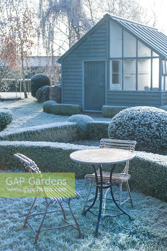 View to garden store and lean to greenhouse. Wrought iron garden table and seats. Yew and box topiary and dwarf hedges on frosty morning in December - The Mill House, Little Sampford, Essex
