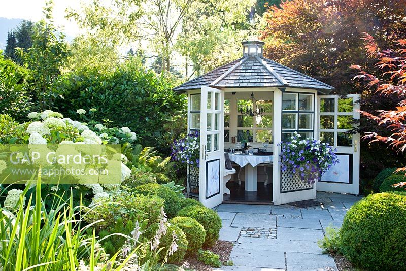White wooden garden pavilion at end of flagstone path. Plants are Buxus, Hosta, Hydrangea arborescens 'Annabell', Petunia Surfinia and Prunus laurocerasus