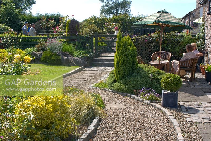 People visiting a National Garden Scheme garden in June with seating area, mixed herbaceous borders and gravel and stone path edged with cobble setts