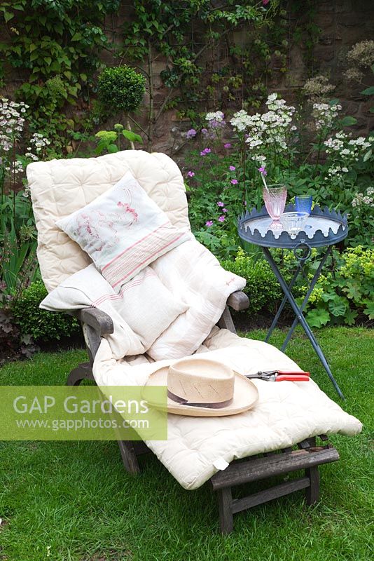 Day bed piled with cushions, with drinks table, straw hat and secaturs in small cottage garden - Garden Neighbours