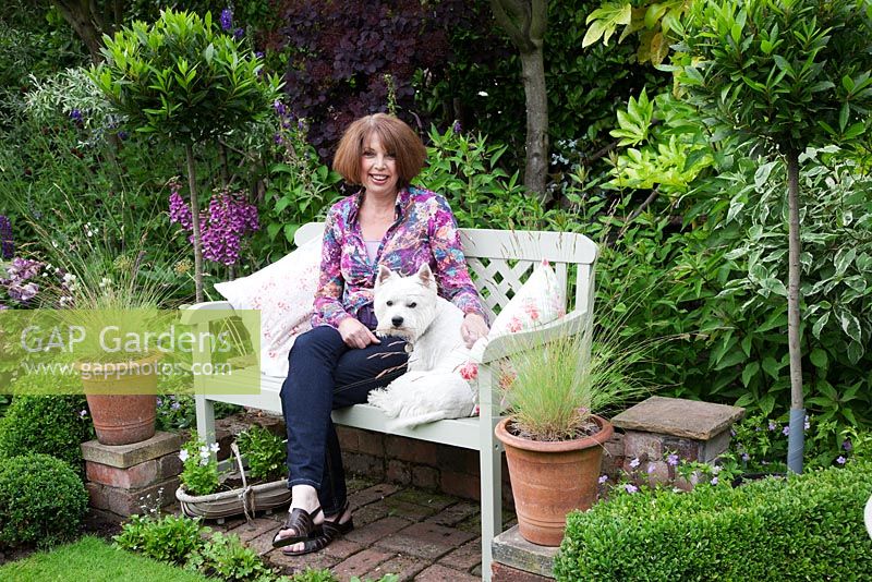Liz, cottage owner sits with Archie on painted bench on brick terrace with Buxus balls and low Buxus hedge, standard Bay trees and potted grasses - Garden Neighbours