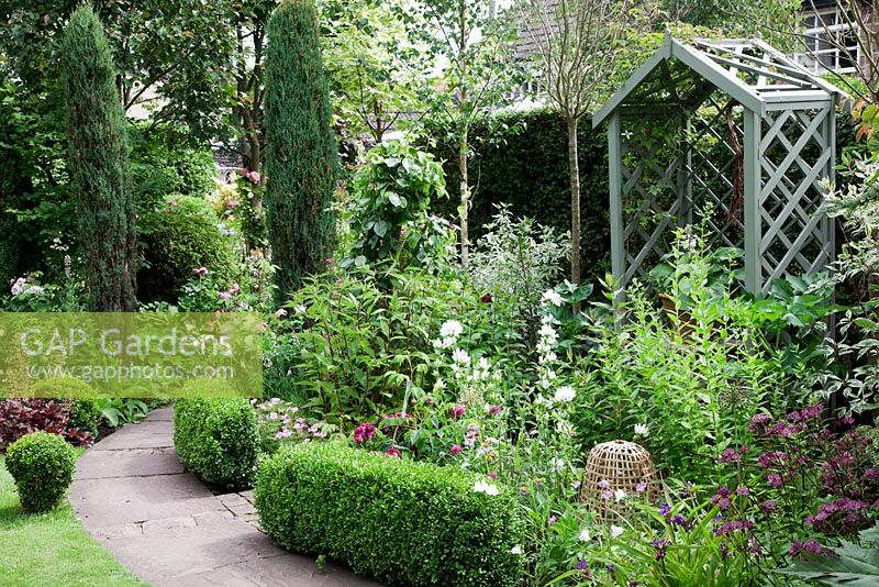 Small cottage garden with stone pathway edged by Buxus low hedging. Two sentinel Juniper  'Skyrocket', wooden bower seat, Astrantia 'Claret', Rosa 'Tuscany Superb', Campanula glomerata and Campanula persicifolia. Garden Neighbours